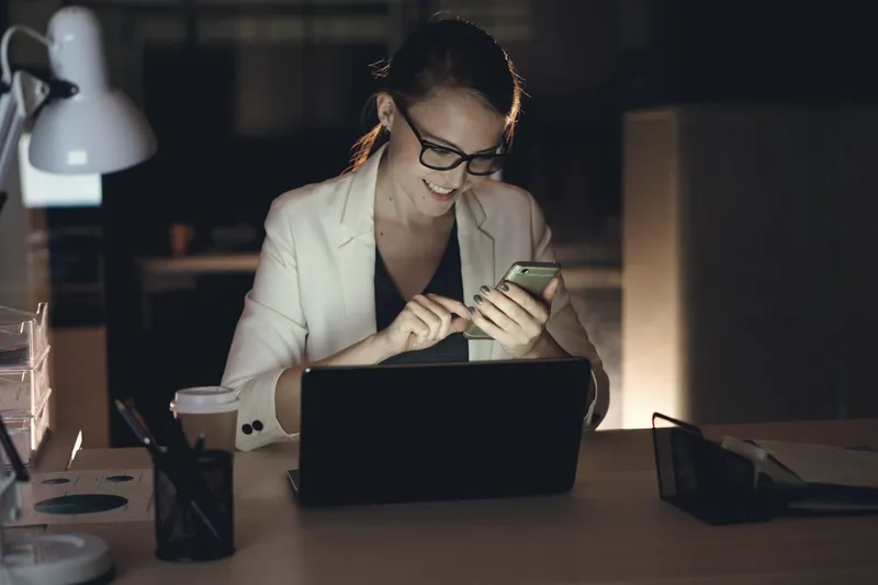 Eine Frau arbeitet abends am Laptop und schaut auf ihr Handy.