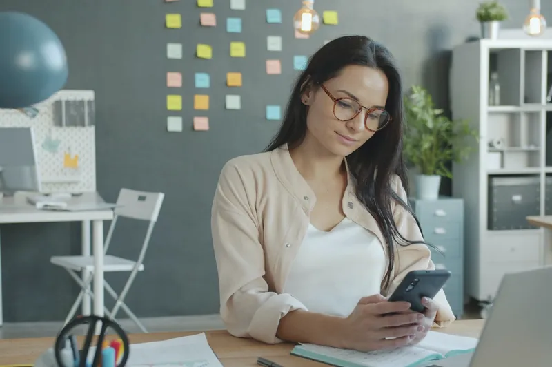 Eine Frau mit Brille sitzt im Büro und schaut auf ihr Handy.